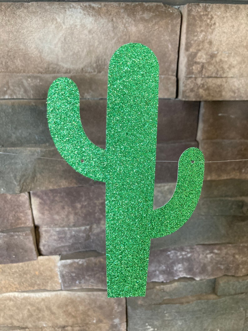 K&ouml;nnte beinhalten: Eine glitzernde, gr&uuml;ne Kaktus-Dekoration. Der Kaktus hat eine strukturierte, funkelnde Oberfl&auml;che und ist vor einem Hintergrund aus gestapelten Steinen aufgeh&auml;ngt. Das Design zeigt die klassische Form eines Saguaro-Kaktus.