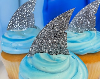 May include: Three cupcakes with blue frosting and silver glitter shark fin toppers. The cupcakes are on a white surface.