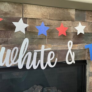 May include: A patriotic banner with red, white, and blue glitter stars strung across a string. The banner also features the words "white & two" in a cursive font. The banner is displayed in front of a stone fireplace.