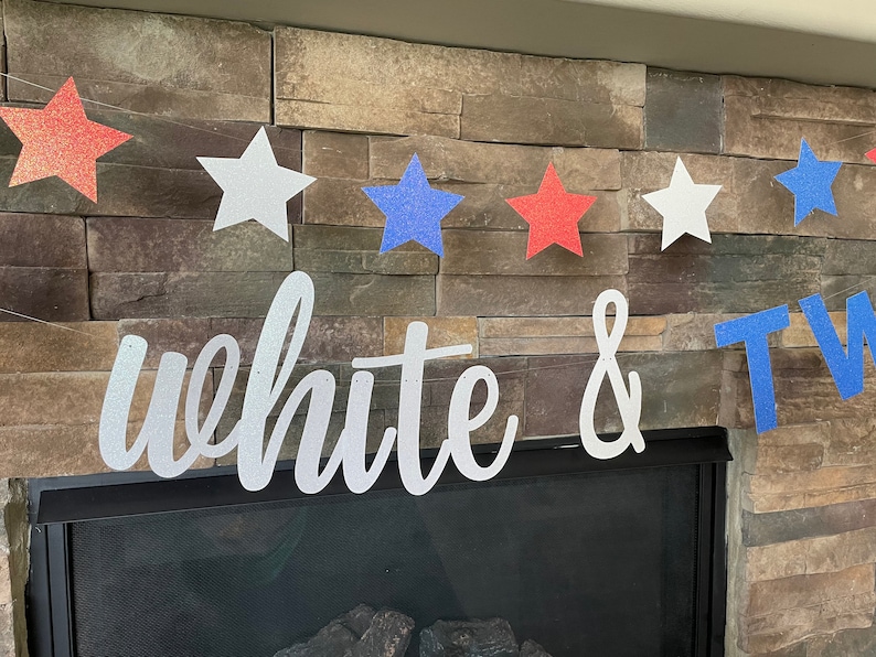 May include: A patriotic banner with red, white, and blue glitter stars strung across a string. The banner also features the words "white & two" in a cursive font. The banner is displayed in front of a stone fireplace.