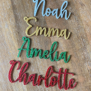 May include: Four glitter-covered names, Noah in silver, Emma in gold, Amelia in green, and Charlotte in red, are arranged in a vertical stack on a wood surface.