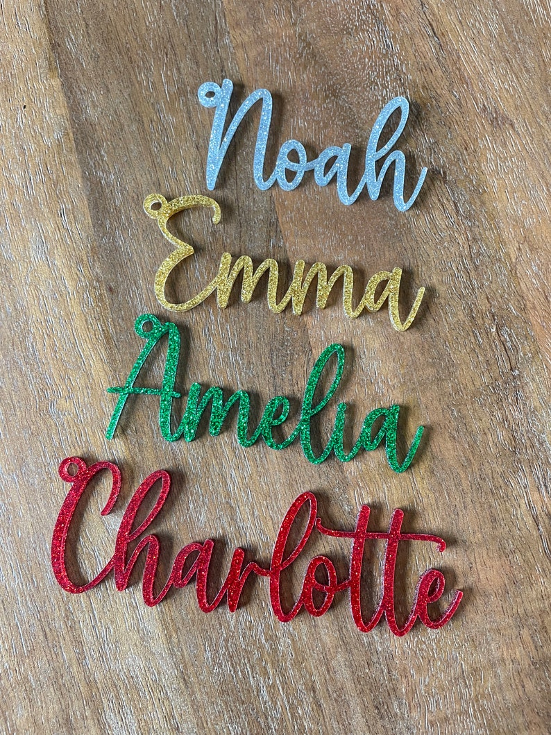 May include: Four glitter-covered names, Noah in silver, Emma in gold, Amelia in green, and Charlotte in red, are arranged in a vertical stack on a wood surface.
