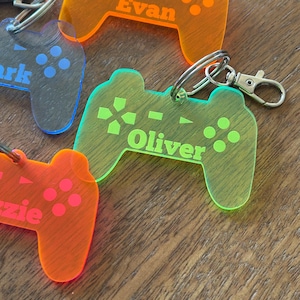 May include: Close-up of several colorful acrylic keychains shaped like video game controllers. Each keychain is a different neon color, with engraved details and a name. The names "Evan", "Oliver", and "Ezzie" are visible.