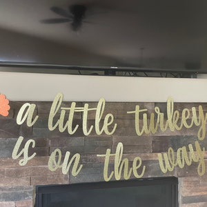 May include: A gold glitter banner with the phrase "a little turkey is on the way" in cursive script. The banner is decorated with two orange and gold turkey cutouts, perfect for a Thanksgiving or baby shower celebration.