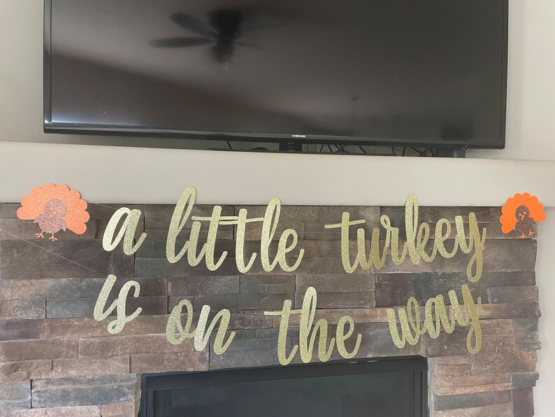 May include: A gold glitter banner with the phrase "a little turkey is on the way" in cursive script. The banner is decorated with two orange and gold turkey cutouts, perfect for a Thanksgiving or baby shower celebration.