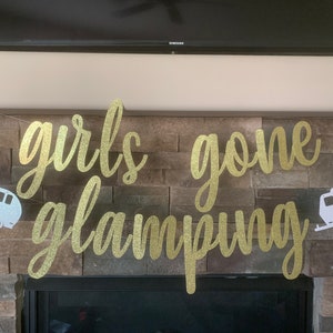 May include: A gold glitter banner with the words "girls gone glamping" in a cursive font. Two white camper trailer cutouts flank the text. The banner is strung across a stone fireplace.