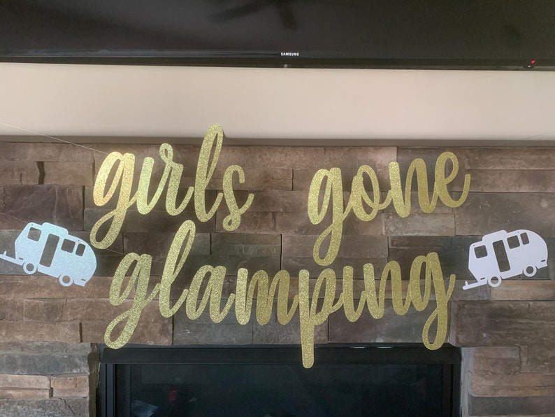 May include: A gold glitter banner with the words "girls gone glamping" in a cursive font. Two white camper trailer cutouts flank the text. The banner is strung across a stone fireplace.