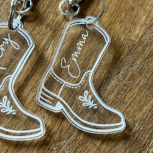 May include: Clear acrylic keychain in the shape of a cowboy boot with the name "Emma" in cursive script. The boot design features white outlines and decorative floral accents, attached to a silver-colored keyring.