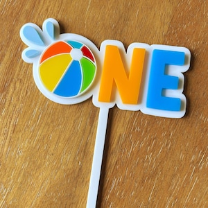 May include: A colorful cake topper with the word "ONE" in orange and blue letters. A beach ball graphic with blue water splashes is on the left. The topper is white with a white stick.