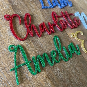 May include: Four names, Liam, Charlotte, Amelia, and a fourth name, are written in glitter on a wooden surface. The names are in different colors: blue, red, green, and gold.