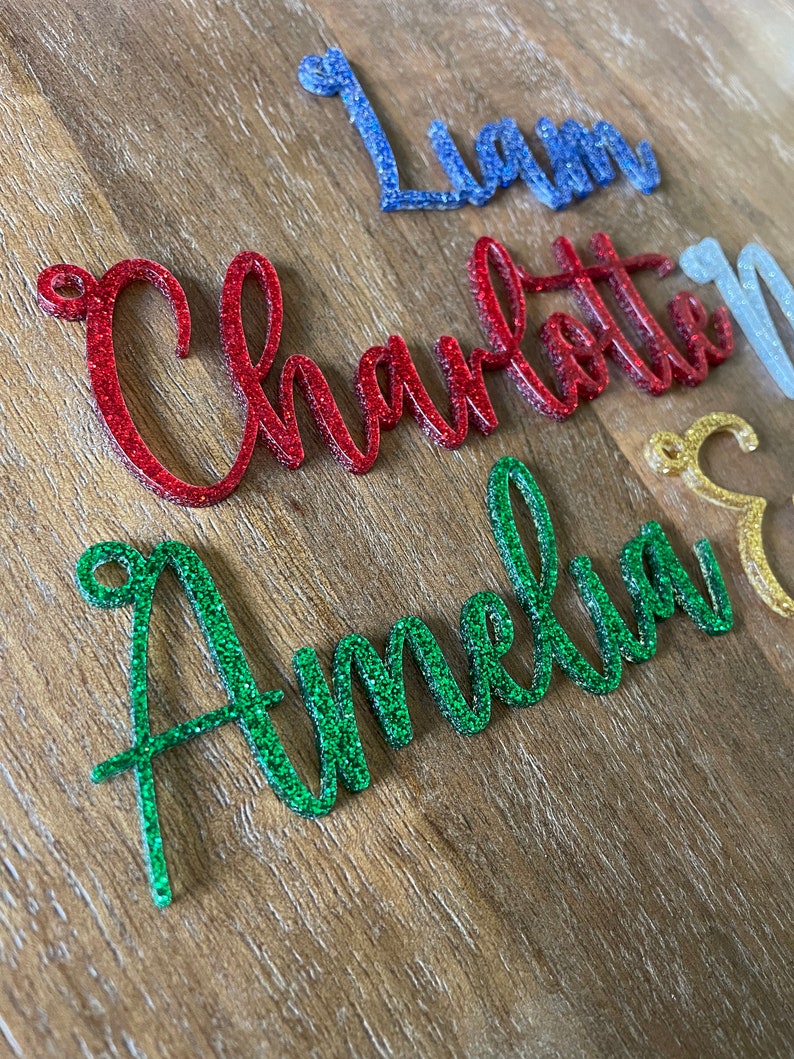 May include: Four names, Liam, Charlotte, Amelia, and a fourth name, are written in glitter on a wooden surface. The names are in different colors: blue, red, green, and gold.