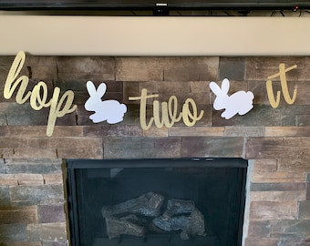 Puede incluir: Una pancarta dorada con purpurina que dice "hop two it" con siluetas de conejos blancos. La pancarta se muestra contra una chimenea de piedra, perfecta para una celebración de segundo cumpleaños.