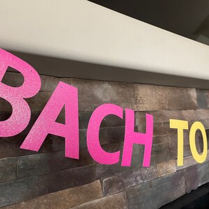 May include: A banner with the words "BACH TO THE" in large, glittery letters. The letters are in bright pink, yellow, and green, and are strung across a stone wall. The banner is likely for a bachelorette party.