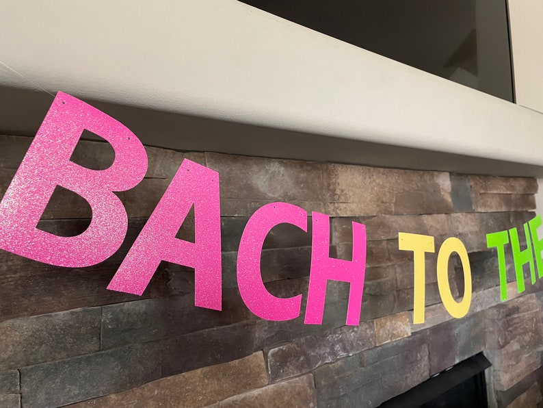 May include: A banner with the words "BACH TO THE" in large, glittery letters. The letters are in bright pink, yellow, and green, and are strung across a stone wall. The banner is likely for a bachelorette party.