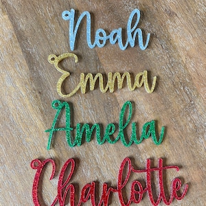 May include: Four glitter-covered names, Noah in silver, Emma in gold, Amelia in green, and Charlotte in red, are arranged vertically on a woodgrain surface.