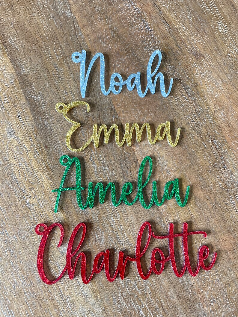 May include: Four glitter-covered names, Noah in silver, Emma in gold, Amelia in green, and Charlotte in red, are arranged vertically on a woodgrain surface.