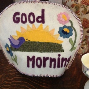 May include: White felt tea cozy with a purple bird, yellow sun, and green grass applique. The text "Good Morning" is embroidered in purple.