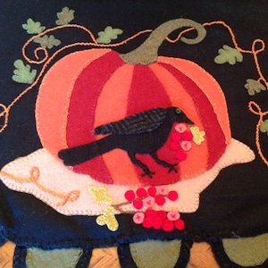 May include: A black felt appliqué of a pumpkin with a crow perched on top. The crow is holding a bunch of red berries in its beak. The pumpkin is surrounded by a vine with green leaves. The background is black felt.