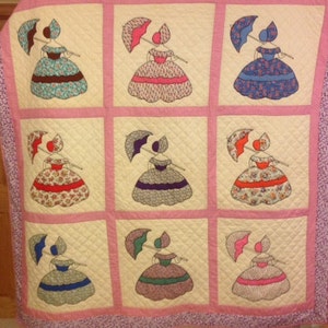Peut inclure: Une courtepointe vintage avec un fond rose et neuf carrés. Chaque carré représente une femme en robe colorée tenant un parapluie. La courtepointe est matelassée avec un motif en losange.