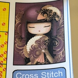 May include: A cross-stitch pattern featuring a stylized doll with closed eyes, adorned with a decorative headpiece and fan. The doll has a purple background with ornate gold and cream-colored accents. The text "Cross Stitch" is visible.
