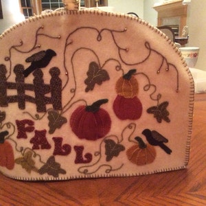 May include: A beige felt tea cozy with an autumn theme. It features a brown fence, black birds, green leaves, and orange and red pumpkins. The word "FALL" is stitched in brown on the front.