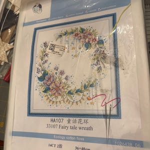 May include: A cross-stitch kit featuring a floral wreath design with pink and blue flowers, hanging ornaments, and the text "Fairy tale wreath." The kit includes cotton fabric, thread, and a needle. The finished size is 14.17 x 15.75 inches.