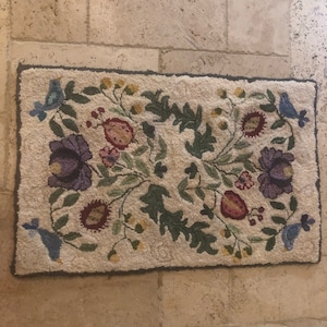 May include: A cream-colored rug with a floral design featuring purple, red, and green flowers, and blue birds. The rug has a dark gray border.