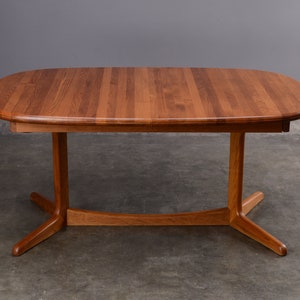 May include: An oval dining table crafted from medium-toned wood with a visible grain pattern. The tabletop features a smooth, rounded edge and is supported by a central pedestal base that branches into four splayed feet connected by a curved stretcher.