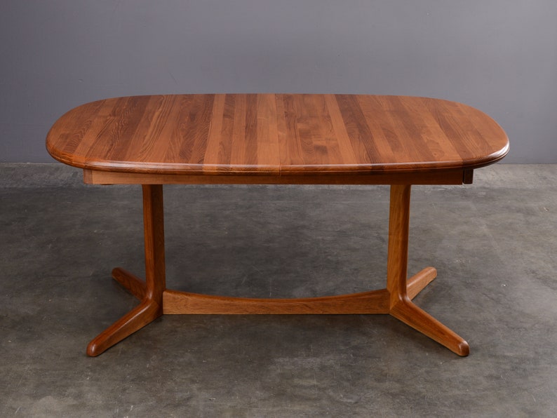 May include: An oval dining table crafted from medium-toned wood with a visible grain pattern. The tabletop features a smooth, rounded edge and is supported by a central pedestal base that branches into four splayed feet connected by a curved stretcher.