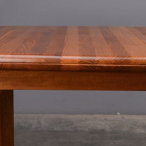 May include: A rectangular wooden dining table featuring a rich, warm brown grain pattern on the surface. The table has a smooth, rounded edge profile and a sturdy wooden leg visible on the left side, showcasing the natural texture of the wood.
