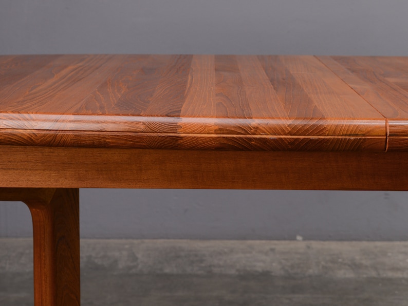 May include: A rectangular wooden dining table featuring a rich, warm brown grain pattern on the surface. The table has a smooth, rounded edge profile and a sturdy wooden leg visible on the left side, showcasing the natural texture of the wood.