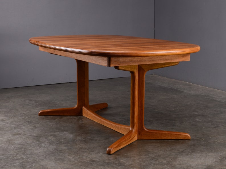 May include: A rectangular wooden dining table with rounded edges and a smooth, warm-toned grain. The table features a central support structure with two vertical pillars connected by a horizontal base, each ending in two splayed feet.
