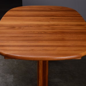 May include: A wooden dining table with a smooth, rounded rectangular top featuring a warm, medium-brown wood grain pattern. The table surface is composed of horizontal planks joined together, supported by a central pedestal base made of matching wood.