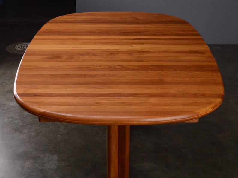 May include: A wooden dining table with a smooth, rounded rectangular top featuring a warm, medium-brown wood grain pattern. The table surface is composed of horizontal planks joined together, supported by a central pedestal base made of matching wood.