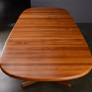 May include: A large oval dining table crafted from warm-toned wood with a visible horizontal grain pattern. The tabletop features a smooth, rounded edge design and is supported by a central pedestal base with extending feet visible underneath.