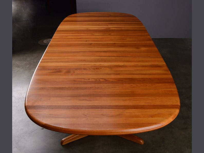 May include: A large oval dining table crafted from warm-toned wood with a visible horizontal grain pattern. The tabletop features a smooth, rounded edge design and is supported by a central pedestal base with extending feet visible underneath.