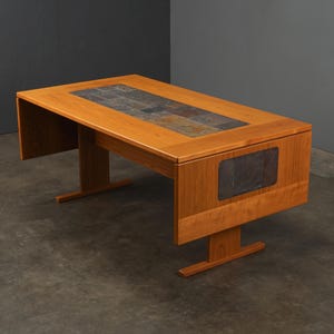 May include: A rectangular wooden table featuring a warm, light brown finish and a central inlay of dark, multi-toned slate tiles. The table has two drop-leaf sides, each accented with a rectangular slate tile inlay, supported by two T-shaped legs.