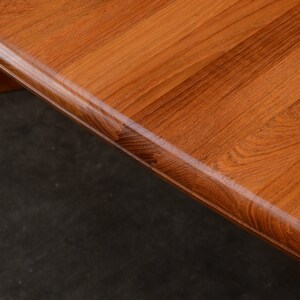 May include: A close-up view of a wooden table surface featuring a warm, medium-brown tone with a prominent, natural wood grain pattern. The edge of the table is rounded and smooth, showcasing the texture and finish of the polished wood material.