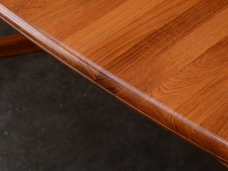 May include: A close-up view of a wooden table surface featuring a warm, medium-brown tone with a prominent, natural wood grain pattern. The edge of the table is rounded and smooth, showcasing the texture and finish of the polished wood material.