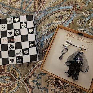 Könnte beinhalten: Eine schwarz-weiß karierte Schachtel mit Halloween-Motiven, darunter Totenköpfe, Herzen und Fledermäuse. Daneben befindet sich eine Schachtel mit einem Paar Ohrringen mit einem gruseligen Thema, darunter eine Skelettbraut und eine Fledermaus.