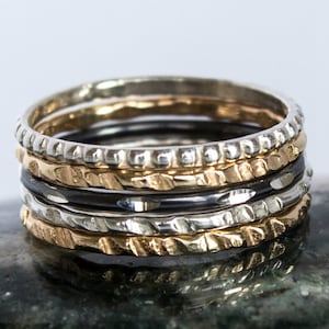 May include: Set of five stacking rings in silver, gold, and black metal. The rings have different textures, including hammered, beaded, and twisted.