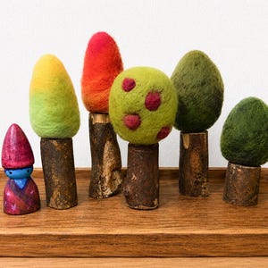 May include: A set of five needle-felted trees with wool tops in yellow, orange, red, and green, mounted on natural wooden branch trunks, alongside a small painted wooden figurine with a purple hat and blue face, all arranged on a wooden tray.