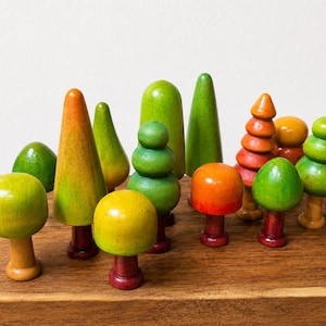May include: A collection of small, hand-painted wooden trees and mushrooms in various shapes and sizes. The pieces feature smooth, rounded forms in vibrant shades of green, yellow, and orange, with distinct brown or red stems for the mushroom figures.