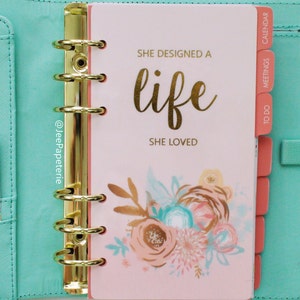 May include: A planner with a light pink cover featuring gold lettering that reads "SHE DESIGNED A LIFE SHE LOVED." The planner has gold rings and coral-colored divider tabs. Floral illustrations in peach, teal, and gold adorn the cover.