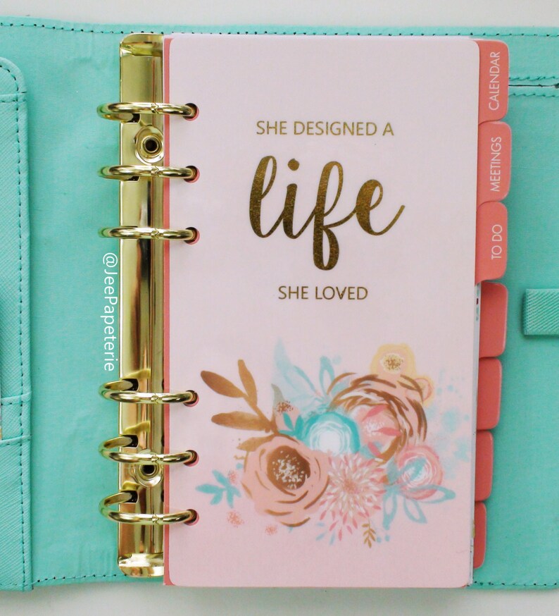 May include: A planner with a light pink cover featuring gold lettering that reads "SHE DESIGNED A LIFE SHE LOVED." The planner has gold rings and coral-colored divider tabs. Floral illustrations in peach, teal, and gold adorn the cover.