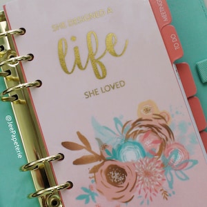 May include: A pink planner with gold accents and a floral design. The cover reads "SHE DESIGNED A LIFE SHE LOVED" in gold lettering. The planner has gold rings and coral-colored divider tabs.