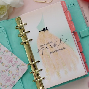 Pode incluir: Um planejador turquesa com an&eacute;is dourados e divis&oacute;rias corais rotuladas como "Calend&aacute;rio", "Reuni&otilde;es" e "A fazer". A capa apresenta uma ilustra&ccedil;&atilde;o de vestido e o texto "Leave a little sparkle wherever you go".