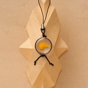 Puede incluir: Un colgante de collar con un dije circular transparente con un diseño de origami amarillo. El dije cuelga de un cordón negro con un detalle metálico plateado. El colgante se muestra contra una escultura de papel geométrica marrón claro.