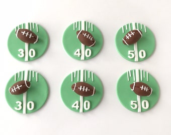 May include: Six green and white fondant cupcake toppers with a football and yard line design. The toppers feature the numbers 30, 40, and 50.