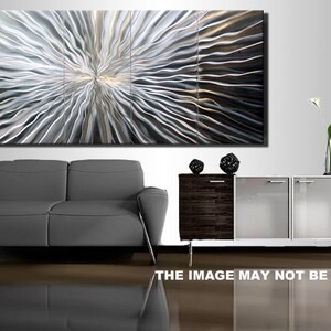 May include: A large abstract metal wall art piece with a silver and gray color scheme. The art is made up of multiple panels and features a swirling, wave-like pattern. The art is displayed on a wall above a gray sofa and a wooden cabinet with glass doors.
